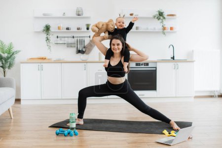 Photo for Full length portrait of joyous lady posing with infant sitting around neck with teddy bear in dining room. Healthy strong mother achieving yoga pose with double weights of daughter and soft companion. - Royalty Free Image