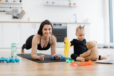 Photo for Joyous caucasian female doing plank workout while focused kid taking care of soft bear on kitchen floor. Pretty brunette mom building endurance before indoor challenge with colorful bands and toys. - Royalty Free Image