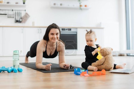 Photo for Joyous caucasian female doing plank workout while focused kid taking care of soft bear on kitchen floor. Pretty brunette mom building endurance before indoor challenge with colorful bands and toys. - Royalty Free Image