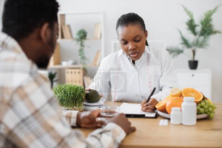 Photo for Focused female specialist writing details about fruit nutrients on clipboard paper while counselling client. Multicultural expert in diet formulating special meals for male patient in clinic. - Royalty Free Image