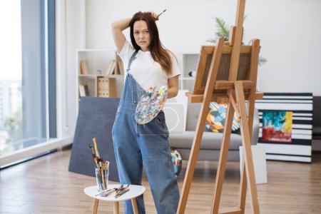Photo for Portrait of beautiful woman in denim overalls posing among artist supplies in spacious home studio. Young female painter with brush and palette honing professional skills while enjoying own space. - Royalty Free Image