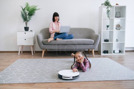 Photo for Focus on well-rounded robot vacuum cleaner being placed on floor of living room by cute child in casual clothes. Stay-at-home mom involving offspring to housekeeping duties while running business. - Royalty Free Image