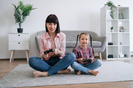Photo for Portrait of cheerful mother and cute daughter sitting cross-legged with input devices in spacious living room. Joyous competitors honing skills in video games while pursuing enjoyable pastime. - Royalty Free Image
