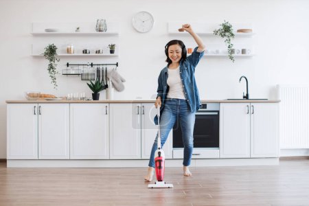 Photo for Smiling vietnamese woman wiggling with uplifting music in headphones while getting floor clean with cordless vacuum. Beautiful housewife entertaining with electric appliance while doing home care. - Royalty Free Image