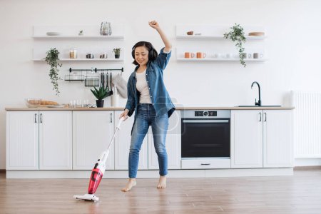 Photo for Smiling vietnamese woman wiggling with uplifting music in headphones while getting floor clean with cordless vacuum. Beautiful housewife entertaining with electric appliance while doing home care. - Royalty Free Image