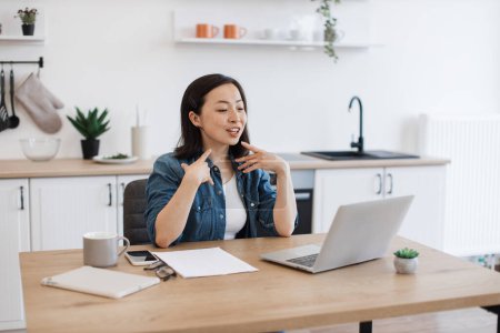 Photo for Smiling asian lady starting video call via internet app on laptop computer in spacious apartment. Delighted entrepreneur introducing herself to online conference attendees on kitchen background. - Royalty Free Image