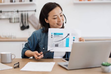 Photo for Cheerful young lady in headset pointing at graphs on paper report while holding online briefing via laptop at home. Efficient female expert interpreting current data for making business predictions. - Royalty Free Image