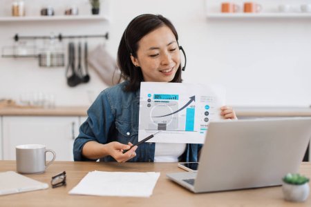 Photo for Cheerful young lady in headset pointing at graphs on paper report while holding online briefing via laptop at home. Efficient female expert interpreting current data for making business predictions. - Royalty Free Image