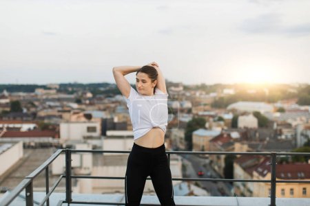 Photo for Attractive brunette female performing overhead triceps stretches while standing near edge of building roof. Young slim woman in cozy outfit increasing range of body motion at end of outdoor training. - Royalty Free Image