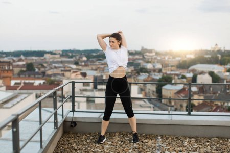 Photo for Attractive brunette female performing overhead triceps stretches while standing near edge of building roof. Young slim woman in cozy outfit increasing range of body motion at end of outdoor training. - Royalty Free Image