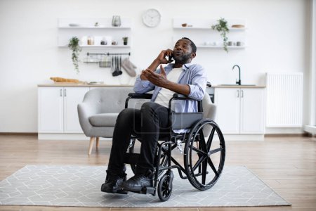 Photo for Full length view of adult person in wheelchair talking on cell phone while using comfort of modern studio flat. Mature african man organising birthday party while getting recovery from illness. - Royalty Free Image