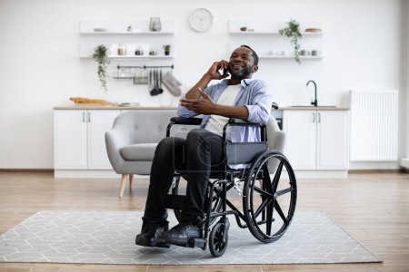Photo for Full length view of adult person in wheelchair talking on cell phone while using comfort of modern studio flat. Mature african man organising birthday party while getting recovery from illness. - Royalty Free Image