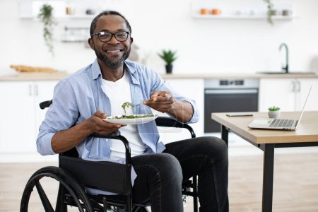 Téléchargez les photos : Salade placée sur un bureau près d'un ordinateur portable par une personne handicapée africaine dans la salle à manger. Happy Business Manager profiter d'aliments sains tout en télécommutant dans le bureau à la maison à midi. - en image libre de droit
