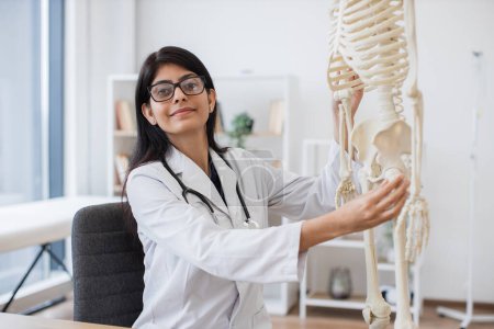 Portrait of positive hospital worker in eyeglasses and lab coat sitting next to model skeleton and looking at camera. Mature hindu woman touching bones on leg and arm for examining disorders.