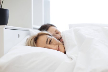 Photo for Pretty caucasian blond woman leaning on soft pillow already awake while her attractive husband still getting shuteye on bed in front of panoramic window. Happy couple spending morning together. - Royalty Free Image