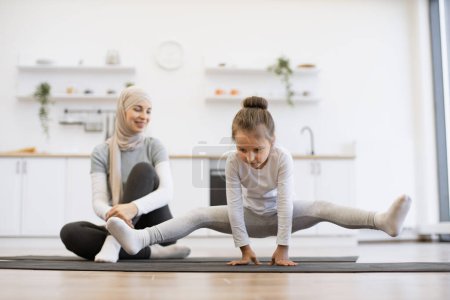 Téléchargez les photos : Jolie fille préscolaire pratiquant le yoga, debout dans l'exercice de la grue, travaillant avec une jeune mère musulmane assise sur un tapis portant des vêtements de sport, à l'intérieur en arrière-plan de la cuisine loft blanc. - en image libre de droit