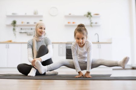 Téléchargez les photos : Jolie fille préscolaire pratiquant le yoga, debout dans l'exercice de la grue, travaillant avec une jeune mère musulmane assise sur un tapis portant des vêtements de sport, à l'intérieur en arrière-plan de la cuisine loft blanc. - en image libre de droit