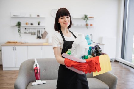 Téléchargez les photos : Portrait d'une jeune femme blanche ouvrière de nettoyage professionnelle tenant un seau pour le lavage avec des détergents sur un fond lumineux de studio de cuisine, espace de copie. - en image libre de droit
