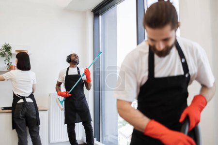 Photo for Team of professional cleaners on background of modern bright kitchen. African male worker washes panoramic windows, having fun while holding mop like guitar, listening to favorite song in headphones. - Royalty Free Image