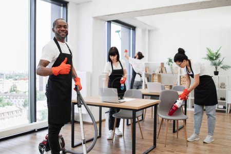 Team of multicultural cleaners wipes tables and gadgets, shelves, washes windows in spacious, bright, modern office. African American man using vacuum cleaner to clean floor from dirt showing thumb up
