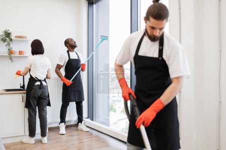 Photo for Focus on positive African American worker cleaning panoramic window glass with mop in modern apartment near multiethnic co-workers. - Royalty Free Image
