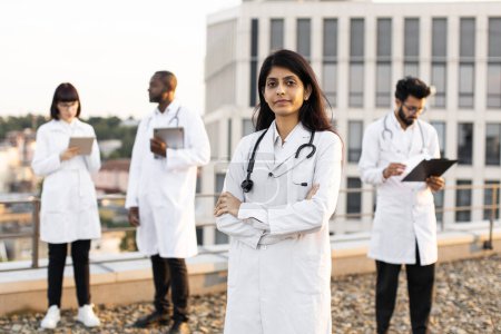 Téléchargez les photos : Portrait d'un jeune médecin indien confiant en blouse blanche posant à la caméra les mains croisées et souriant. Collèges internationaux de nationalité mixte se tenant derrière et opérant gadgets modernes. - en image libre de droit