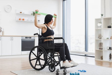 Vue complète de la femme caucasienne en fauteuil roulant étirant bandes tout en exerçant avec mobilité réduite à l'intérieur. Adulte souriant utilisant une activité d'intensité modérée pendant l'entraînement à la maison dans la cuisine.