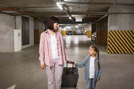 Téléchargez les photos : Jeune femme avec enfant tenant une valise de voyage comme recherche de voiture avant d'embarquer sur le voyage. Mère caucasienne et petite fille vêtus de vêtements décontractés marchant dans un parking souterrain. - en image libre de droit