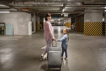 Téléchargez les photos : Jeune femme avec son enfant serrant une valise de voyage à la recherche d'un véhicule avant de commencer le voyage. Joyeux maman et petite fille, vêtus de vêtements informels, se promener dans l'aire de stationnement souterraine. - en image libre de droit