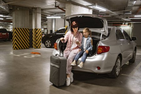 Téléchargez les photos : Mère caucasienne et petite fille assis dans le coffre de la voiture moderne dans le parking souterrain. Jeune femme avec enfant tenant une valise de voyage en utilisant le téléphone pour planifier un voyage de vacances. - en image libre de droit