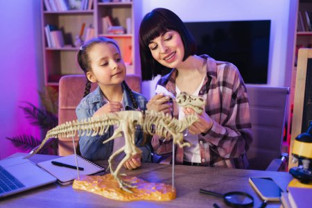 Caring woman and small kid girl improving knowledge at evening home. Caucasian mother assemble skeleton of dinosaur with smart cute daughter while making model of tyrannosaurus using glue.
