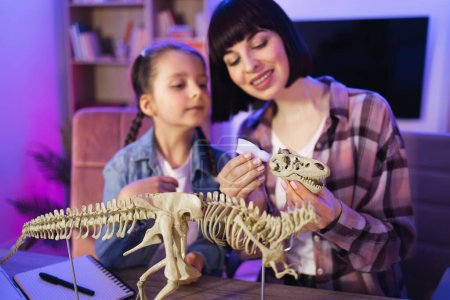 Fürsorgliche Frau und kleines Mädchen verbessern ihr Wissen zu Hause am Abend. Kaukasische Mutter baut Dinosaurier-Skelett mit smarter Tochter zusammen, während sie Tyrannosaurus-Modell mit Klebstoff herstellt.