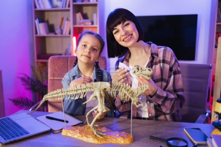 Caring woman and small kid girl improving knowledge at evening home. Caucasian mother assemble skeleton of dinosaur with smart cute daughter while making model of tyrannosaurus using glue.