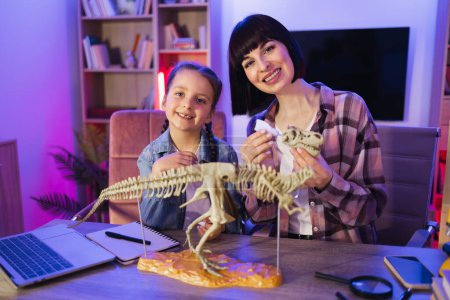 Caucasian mother assemble skeleton of dinosaur with smart cute daughter while making model of tyrannosaurus using glue. Caring woman and small kid girl improving knowledge at evening home.