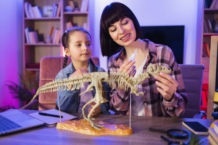 Téléchargez les photos : Mère caucasienne assembler squelette de dinosaure avec fille mignonne intelligente tout en faisant modèle de tyrannosaure à l'aide de colle. Femme bienveillante et petite fille améliorant les connaissances à la maison du soir. - en image libre de droit