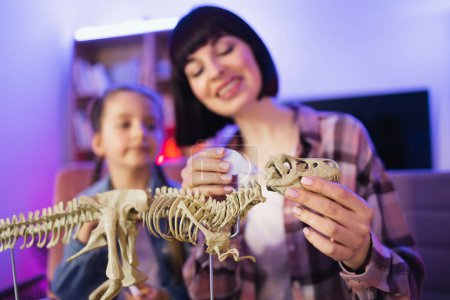Téléchargez les photos : Mère caucasienne assembler squelette de dinosaure avec fille mignonne intelligente tout en faisant modèle de tyrannosaure à l'aide de colle. Femme bienveillante et petite fille améliorant les connaissances à la maison du soir. - en image libre de droit