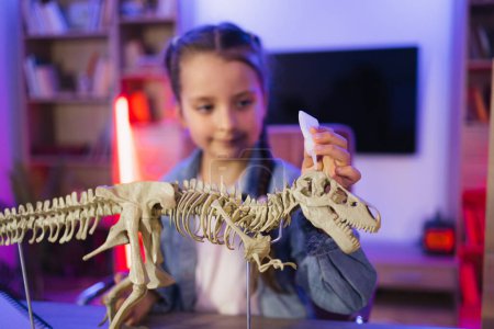 Petite fille scientifique caucasienne de six ans en vêtements décontractés assis à la table examiner le squelette de dinosaure. Joyeux enfant préscolaire collant os faisant modèle de tyrannosaure à la maison du soir.