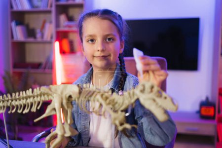 Téléchargez les photos : Joyeux enfant d'âge préscolaire collant os faisant modèle de tyrannosaure à la maison du soir. Petite fille scientifique caucasienne de six ans en vêtements décontractés assis à la table examiner le squelette de dinosaure. - en image libre de droit