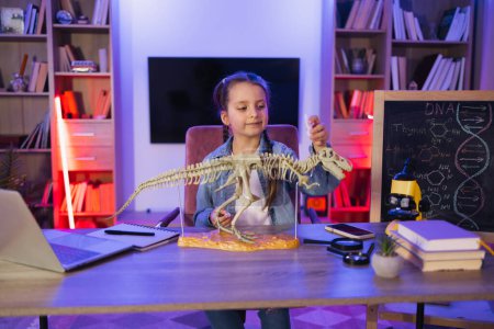 Joyeux enfant d'âge préscolaire collant os faisant modèle de tyrannosaure à la maison du soir. Petite fille scientifique caucasienne de six ans en vêtements décontractés assis à la table examiner le squelette de dinosaure.