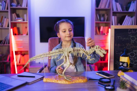 Focused Caucasian little girl study fossil prehistoric animals in evening at living room. Happy preschool child gluing bones making model of tyrannosaurus at evening home.