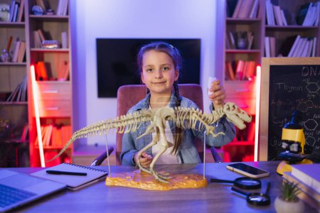 Téléchargez les photos : Petite fille caucasienne ciblée étudier les animaux fossiles préhistoriques en soirée dans le salon. Joyeux enfant préscolaire collant os faisant modèle de tyrannosaure à la maison du soir. - en image libre de droit
