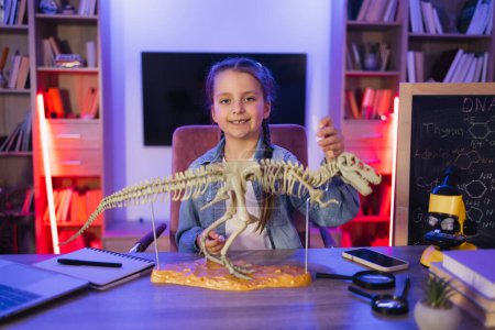 Focused Caucasian little girl study fossil prehistoric animals in evening at living room. Happy preschool child gluing bones making model of tyrannosaurus at evening home.