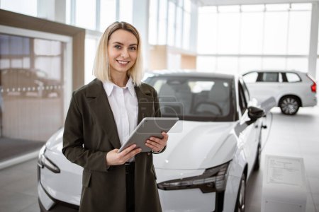 Professional salesperson working in bright and spacious car dealership. Attractive young woman standing in front of car with digital tablet. Beautiful woman in suit in front of luxury vehicle.