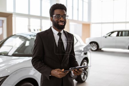 Portrait of confident Caucasian man, salesman in suit holding clipboard in bright modern car service. Young successful manager on background of rows of new car dealership crossovers.
