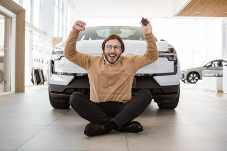 Happy bearded man sitting at lotus pose near new bought car and holding keys in car dealership. Successful buyer in casual outfit rejoices in a new car. Trading concept.