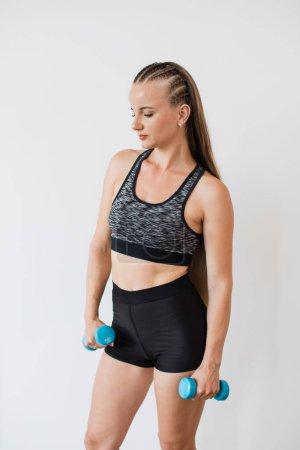 A focused young woman in sports attire performs an exercise routine with blue dumbbells against a white background, exemplifying health and fitness.