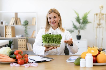 Experienced dietitian adding different vitamins, minerals and nutrients to eating. Charming Caucasian woman in doctors coat with microgreens smiling in consulting room of private clinic.