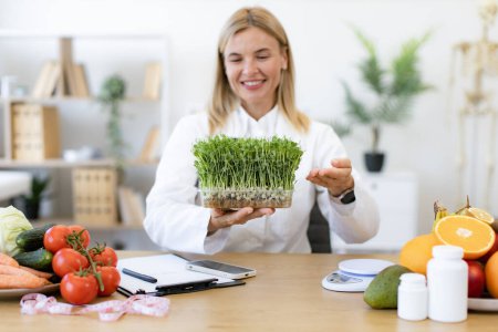 Experienced dietitian adding different vitamins, minerals and nutrients to eating. Charming Caucasian woman in doctors coat with microgreens smiling in consulting room of private clinic.