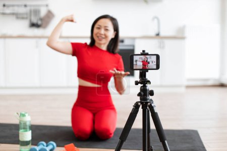 Mujer en ropa deportiva roja grabación de vídeo de fitness en casa. Ella está flexionando los músculos y demostrando ejercicios con equipos de fitness como mancuernas.