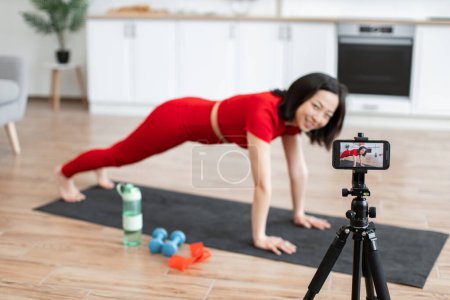 Woman in red activewear filming home workout video in modern kitchen doing yoga exercise planks on mat. Fitness equipment includes water bottle, dumbbells, resistance band.